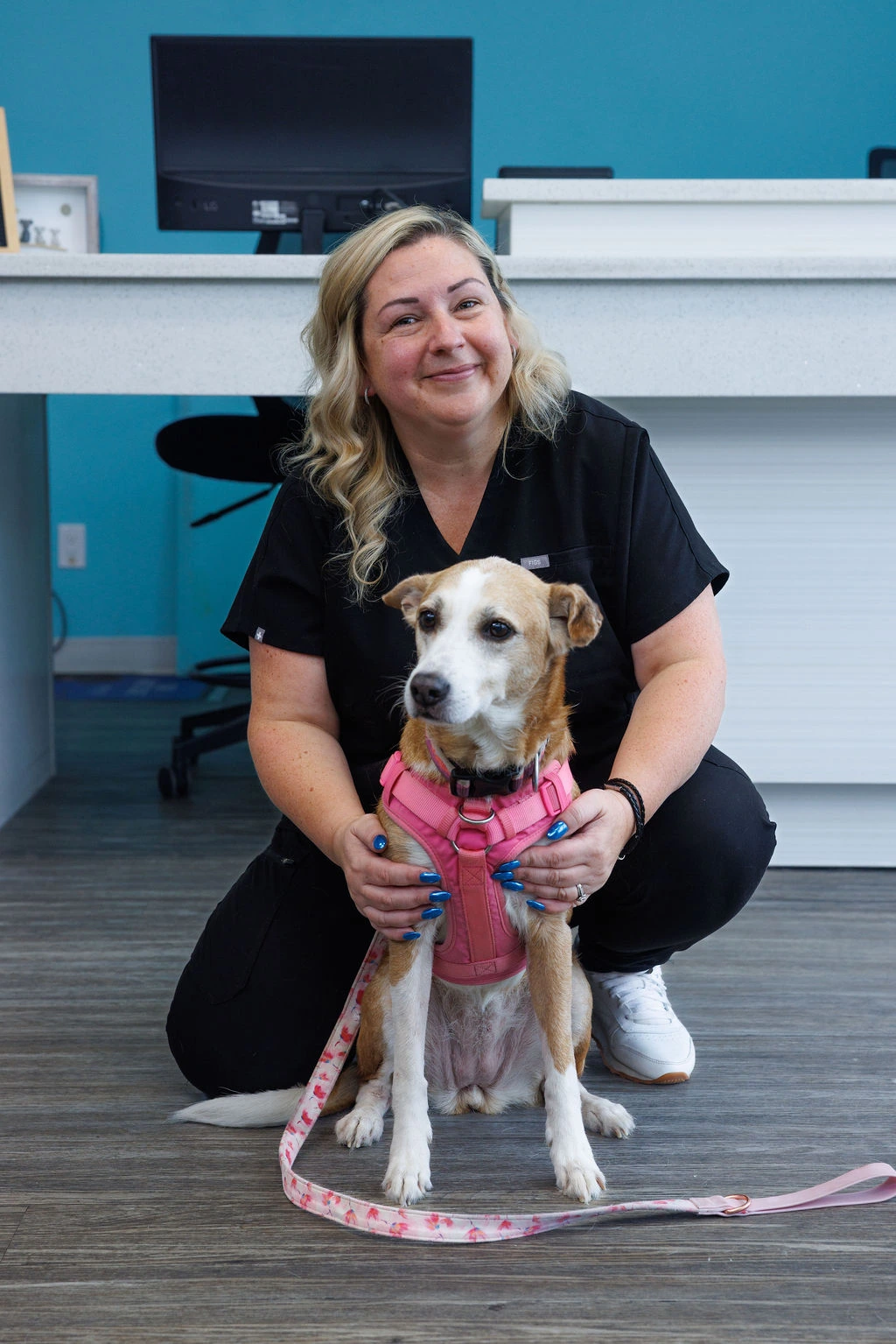 Wendy crouching behind a tan-and-white dog in a pink harness